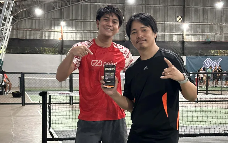 Two men standing on an indoor pickleball court, one wearing a red shirt and pointing at a smartphone held by the other man in a black shirt making a hand gesture. happy to be running their session with the fairplay pickleball app