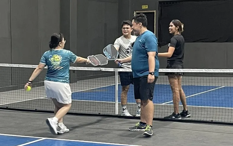 Four people on an indoor pickleball court exchanging paddles over the net, with one person holding a ball. a session run using fairplay pickleball app