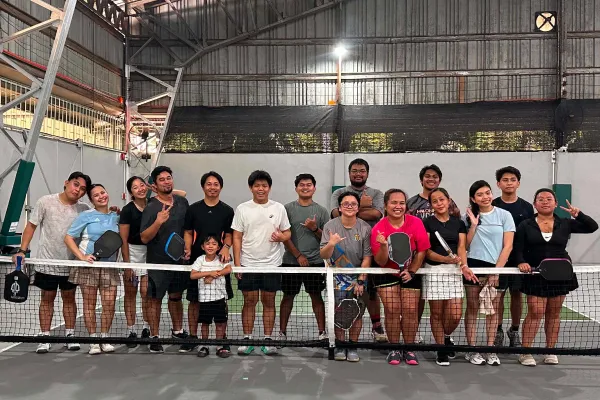 Group of people standing on an indoor pickleball court behind the net, some holding paddles and smiling at the camera. just finished a session that was run using fairplay pickleball app