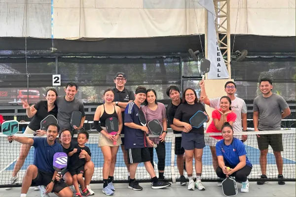 Group of thirteen adults and one child posing with pickleball paddles on an indoor court behind the net. just finished a session that was run using fairplay pickleball app