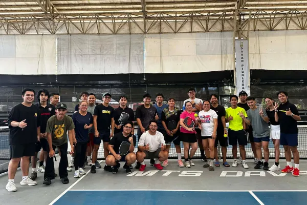 Group of 21 men and women posing with pickleball paddles on an indoor pickleball court under a metal roof. just finished a session that was run using fairplay pickleball app