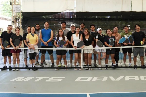 Group of 15 people standing behind a pickleball net on an indoor court holding paddles and smiling, game session powered by fairplay pickleball