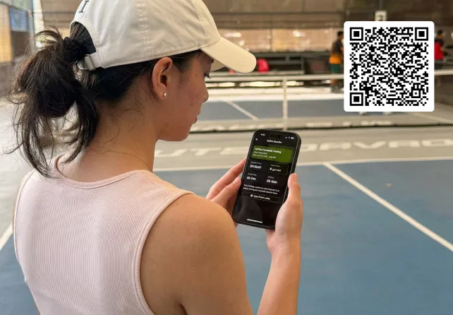 Woman in a white cap viewing a fairplay pickleball app on her phone beside an indoor pickleball court.