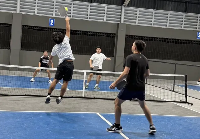 Four young men playing doubles in a pickleball court, one player jumping to hit the pickleball with a paddle.