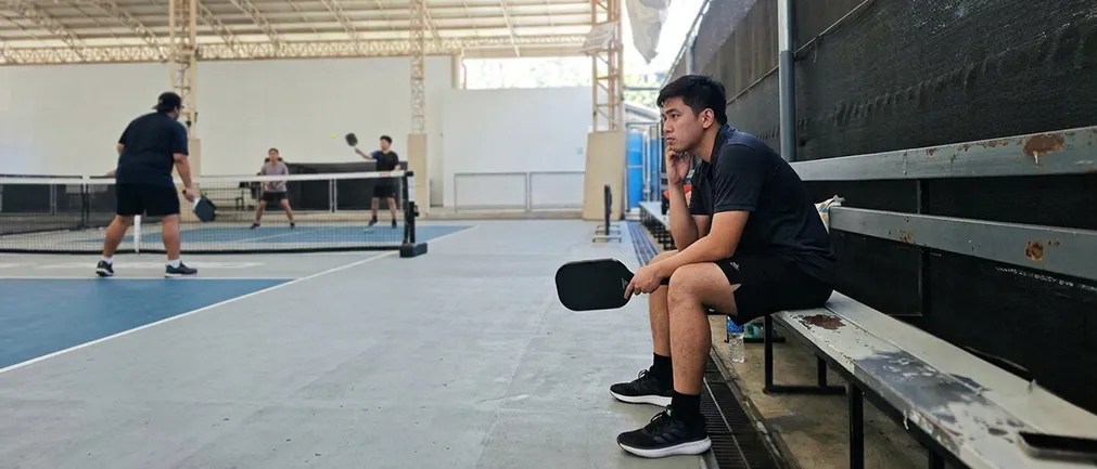 A young man in black sportswear sitting on a bench holding a pickleball paddle, watching a pickleball game in progress on an indoor court.